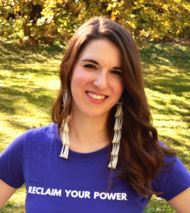 Kirby is wearing a purple t-shirt that says "Reclaim Your Power", long beaded earrings, and is in front of a backdrop of a green grassy yard and trees 