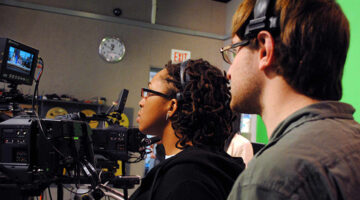 Two students watch camera monitors in studio