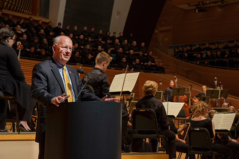 President Clif Smart stands at podium with orchestra on stage