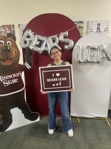 A student standing in front of a maroon & white backdrop, holding a sign that reads "I love Bears Lead."