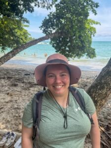 Clara Dickinson smiles while standing on a tropical beach, wearing a pink sunhat, green T-shirt, and a backpack. She is surrounded by trees with ocean waves and swimmers visible in the background.