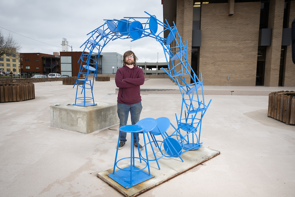 Student standing amid his steel sculpture comprised of 19 objects that resemble metal stools painted blue.