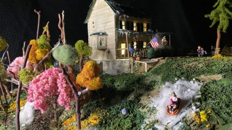 A small model house with whimsical trees and human figures in the yard