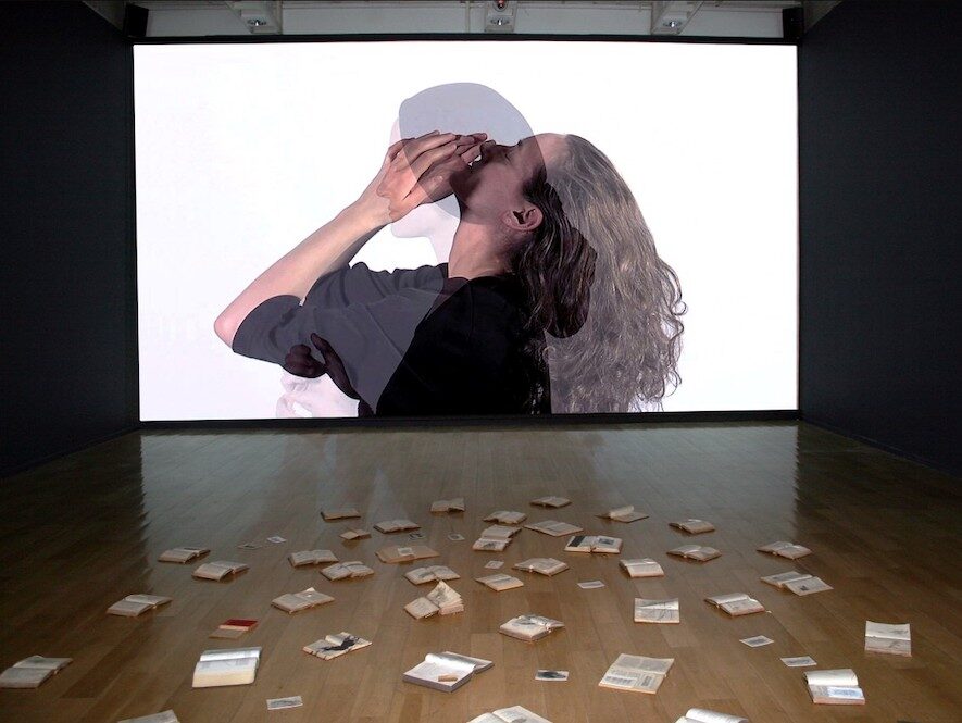 A large screen projecting overlapping side profiles of two women, with open books on the floor below the screen