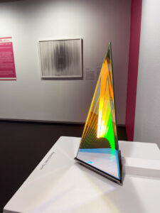 A glass prism sculpture with colorful beams of light sits on a pedastal in front of a framed art piece hanging on a wall.