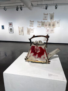 A grey and red ceramic teapot on a pedestal, in front of other artwork hanging on a white wall