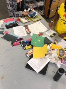 A table covered in paint tubes, colorful paper, and other art supplies. The hands and torsos of two students working at the table are also in frame.