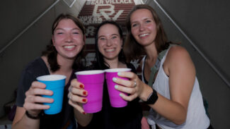 Three females toast with bright colored plastic cups in front of stairs at Plaster Stadium
