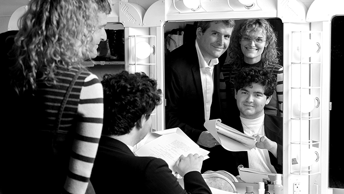A group of individuals gather around a dressing room mirror, reviewing scripts and preparing for a performance.