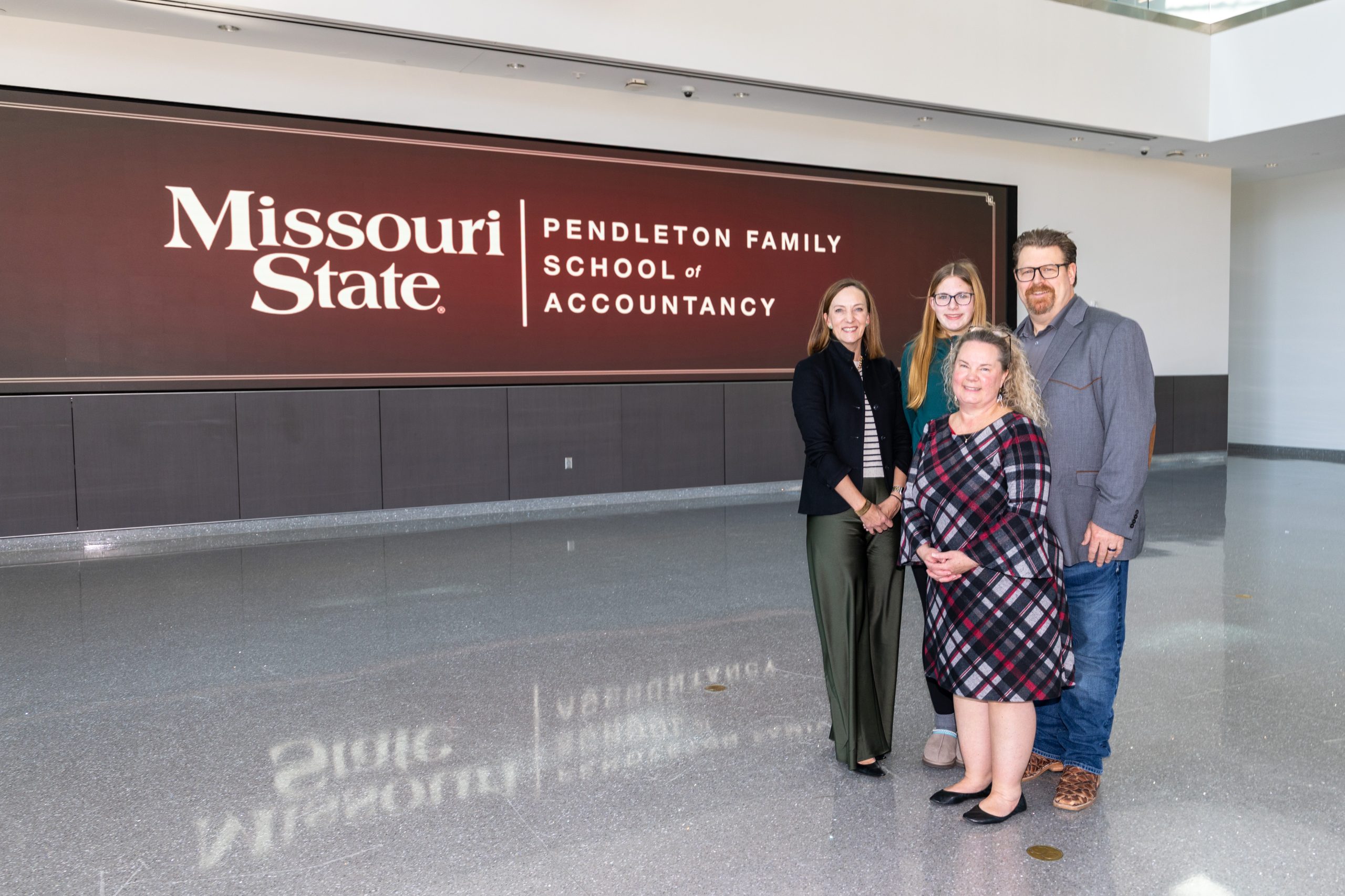 Four smiling people (3 women, 1 man) in business casual attire pose before a large brown sign for the "Missouri State | Pendleton Family School of Accountancy". They stand on a reflective grey floor indoors.