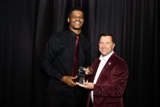 Black man in black shirt/red tie & White man in maroon jacket/white shirt smile, holding a clear athletics award plaque against black drapes.
