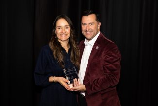 Smiling woman in dark blue and man in maroon jacket hold a clear athletics award plaque against a black backdrop. The plaque shows text including 'ANDREINA "ANDI" ROJAS' and 'MISSOURI STATE UNIVERSITY'.