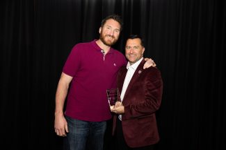 Two smiling men against a black backdrop. The taller man on the left wears a magenta polo and jeans, arm around the shorter man on the right, who wears a maroon corduroy blazer, white shirt, and holds a clear athletics award plaque.