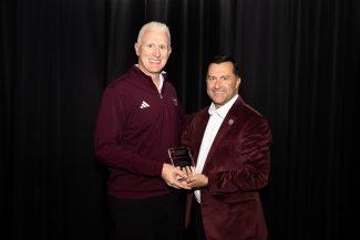 Two smiling men, one in a maroon athletic top & the other in a maroon velvet blazer, jointly hold a clear award plaque. Dark background.