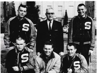 Black & white group photo. An older man in a suit stands with five athletic young men in dark letterman jackets with light sleeves, marked "S STATE." Some hold golf clubs. They are outside a building.