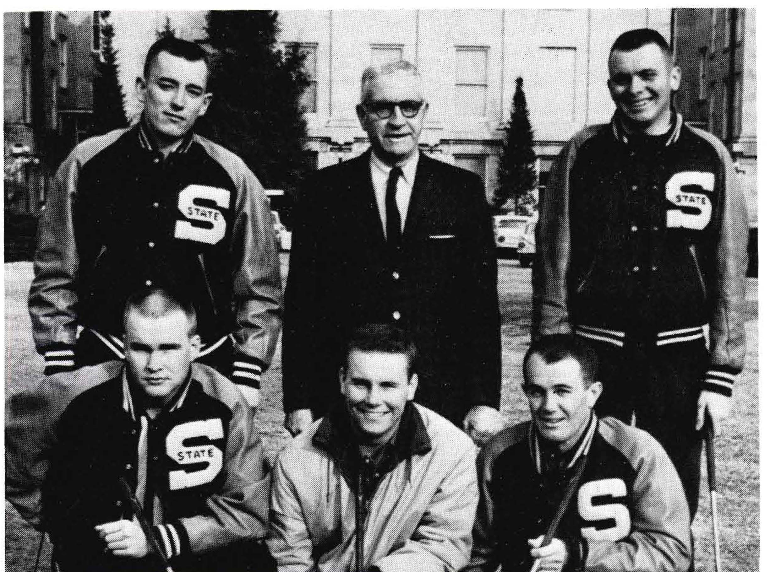 Black and white photo of a 6-man golf team. An older coach in a suit stands behind 5 young men in "S STATE" letterman jackets, holding golf clubs. Outdoors with a building in the background.