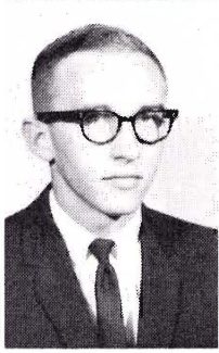 Grainy B&W photo of a young man with short hair, dark-rimmed glasses, a dark suit, white shirt, and tie, looking slightly right.