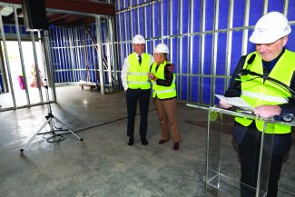 Image of Brent Dunn smiling while hugging Beverly Miller Keltner listening to Jim Wilson deliver remarks in a construction site. All are wearing hardhats and construction vests.