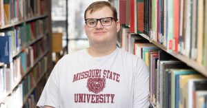 In between two rows of shelves, Daniel Paige leans against a shelf of books at Meyer Library.
