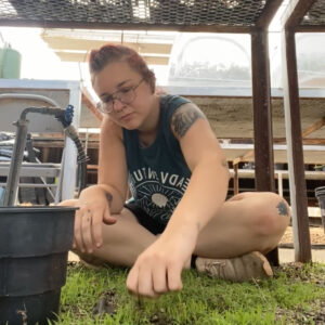 Abilene sits cross-legged next to a water faucet in a greenhouse.