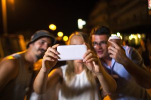 Girl and two male friends taking selfie with smart phone outdoor at night. Blurred street lights in background, focus on the cell