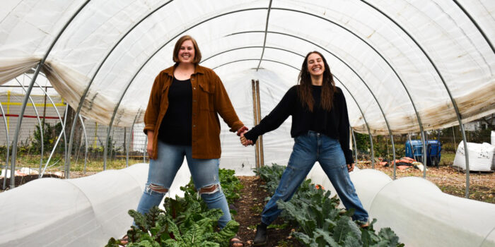 Amanda and Anneliese hold hands and smile as they stand in a garden greenhouse