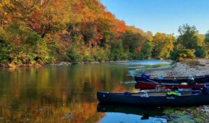 Canoes rest on the shore of a peaceful stream, autumn trees fill the background