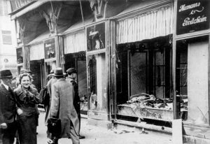 Photo of store front with broken windows and people walking by during Kristallnacht