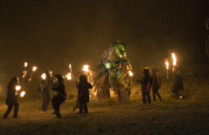 People holding lit torches standing in a circle around a figure made of straw at an Imbolc festival