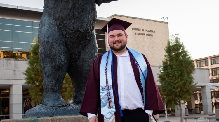 Faron Haase in his graduation attire standing in front of the Bear statue at Plaster Student Union.