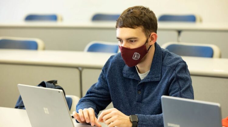 A masked student takes notes on a laptop during class.