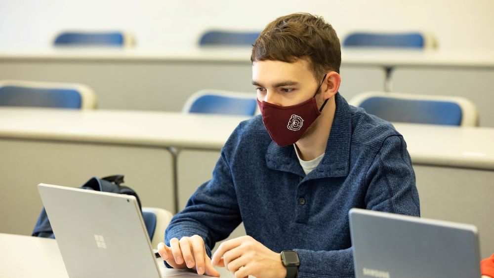 A masked student takes notes on a laptop during class.