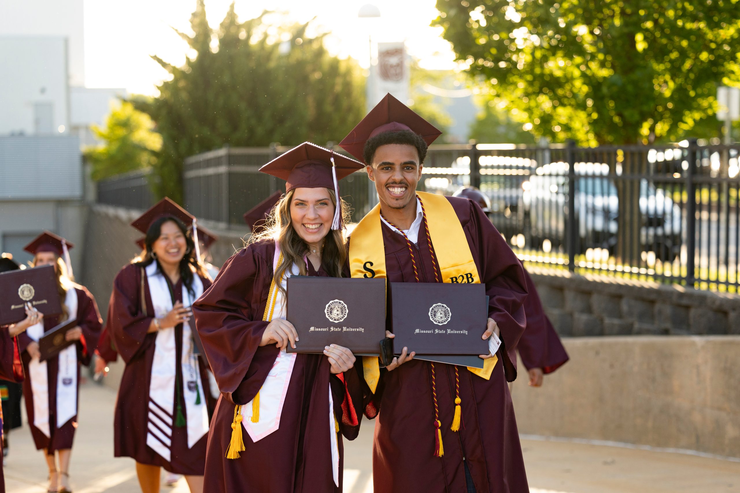 Image of Two people wearing caps and gowns and holding diplomas smile at the camera after graduating from Missouri State