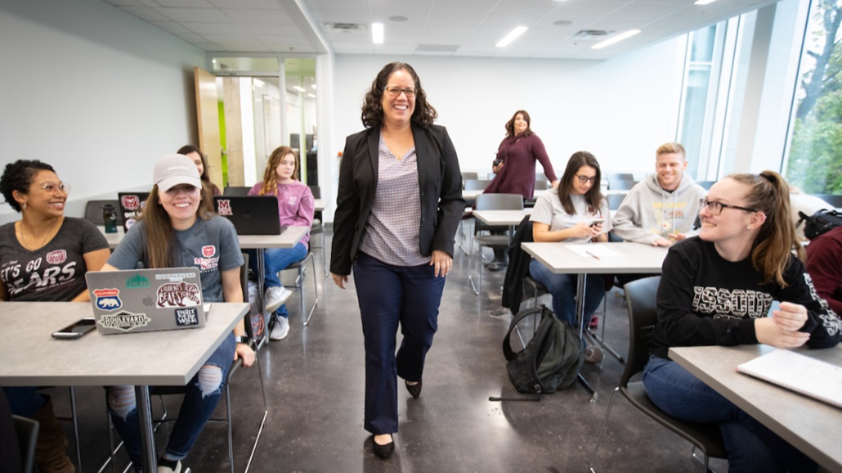 Dr. Liza Cobos passes through aisles of smiling students in classroom.
