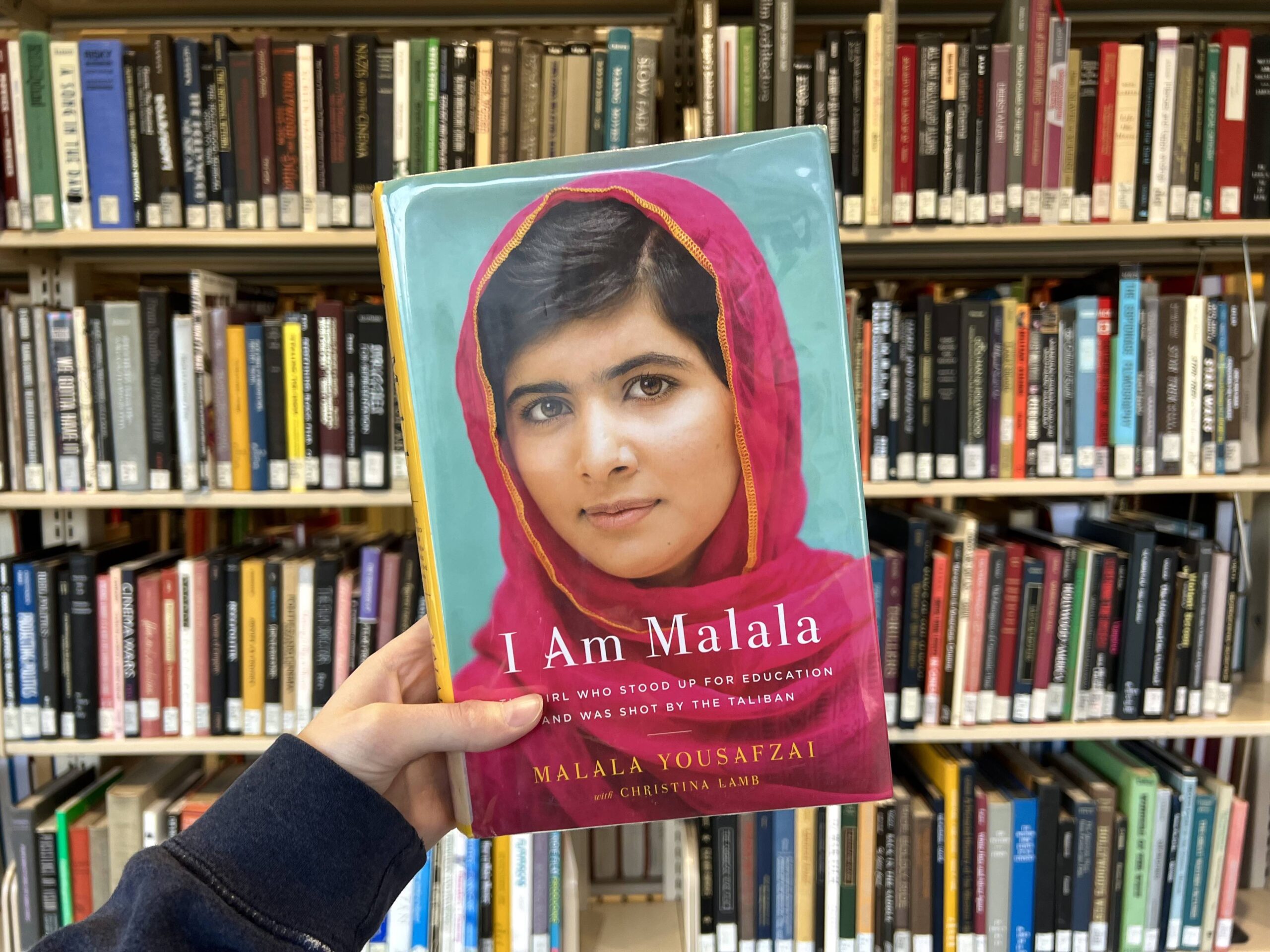 Person holding the book, "I Am Malala," in front of a bookcase.
