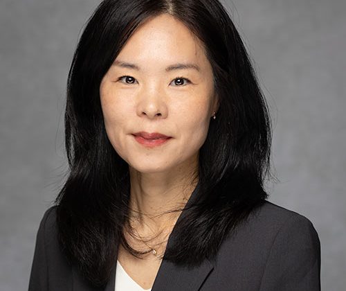 A person with long dark hair wearing a dark blazer over a white top, posed against a neutral gray background.