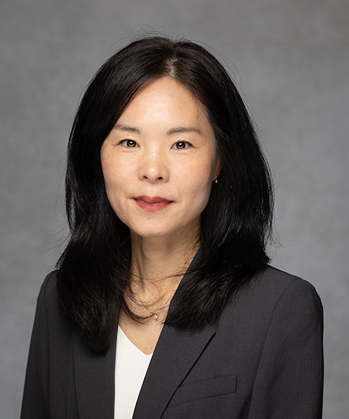 A person with long dark hair wearing a dark blazer over a white top, posed against a neutral gray background.