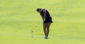 Helena sends dirt flying on the golf course as she swings her club and sends the ball forward.