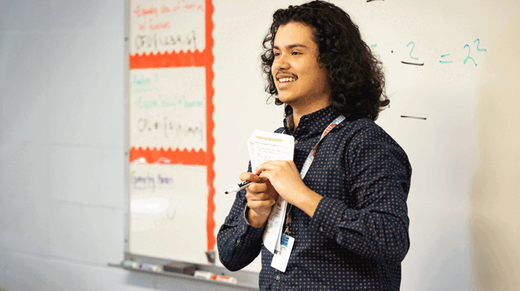 Joseeduardo Granados Rodriguez smiles while standing in front of a whiteboard at the front of a classroom.