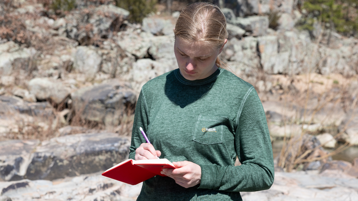 Student jots notes in notebook while standing by river.