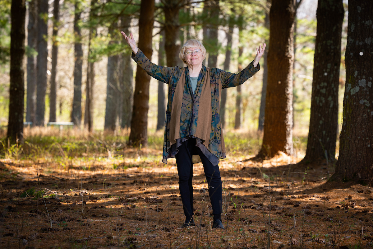 Dr. Carol Miller proudly stands in dappled light, under a beautiful evergreen forest.
