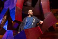 Dr. Senger looks thoughtfully to his left as he leans against a red geometric structure.