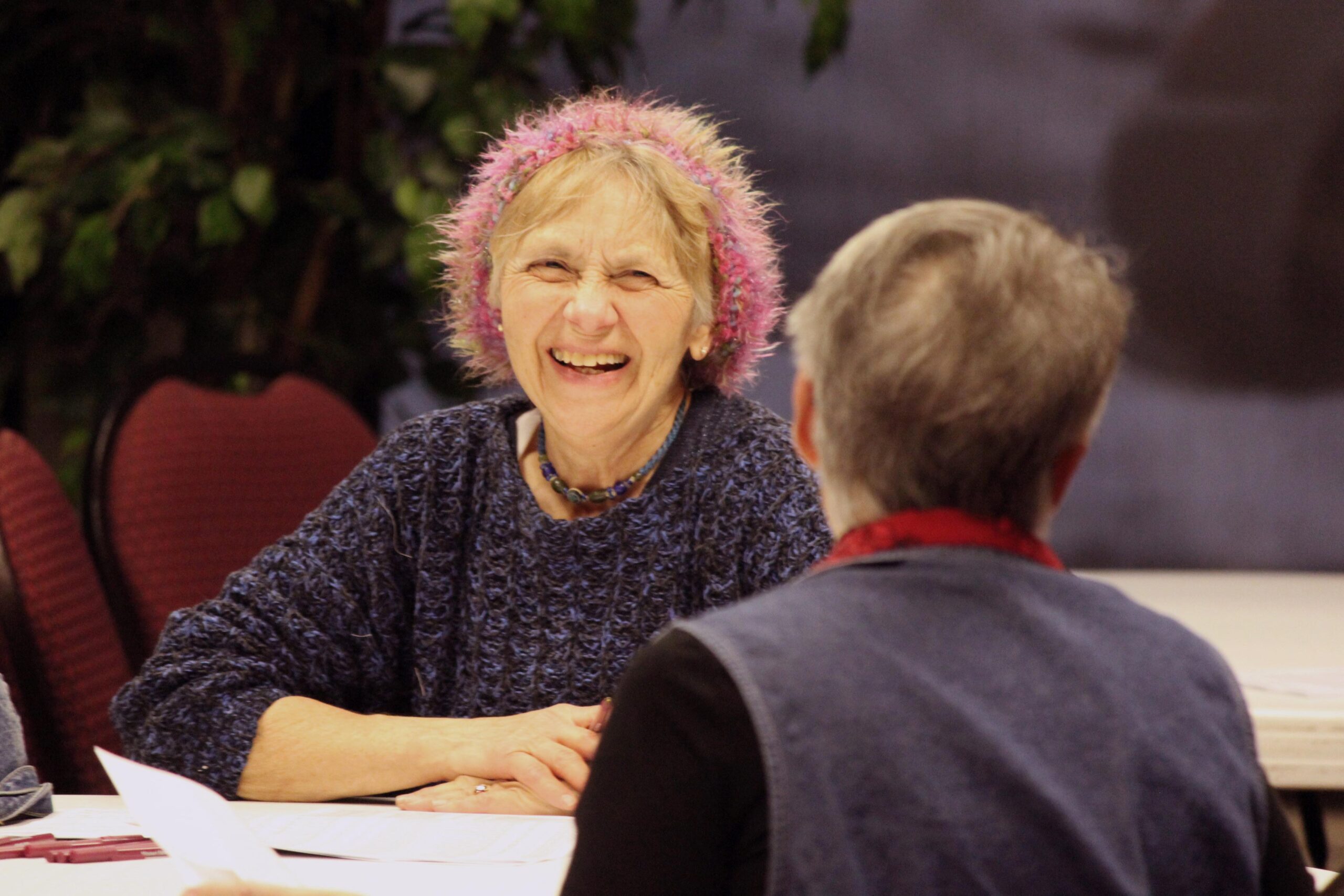 Smiling senior citizen in a pink hat chats across the table with another woman at MSU 62's registration event. 