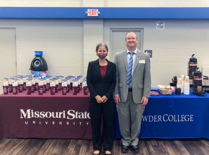 A table draped in MSU maroon and topped with MSU cups sits next to a table draped in Crowder College blue, topped with snacks and coffee. Melissa Warren stands in front of the MSU table. Larry Nichols stands in front of the Crowder College table. 