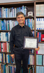 Dr. Ismet Anitsal infront of book shelves holding an award