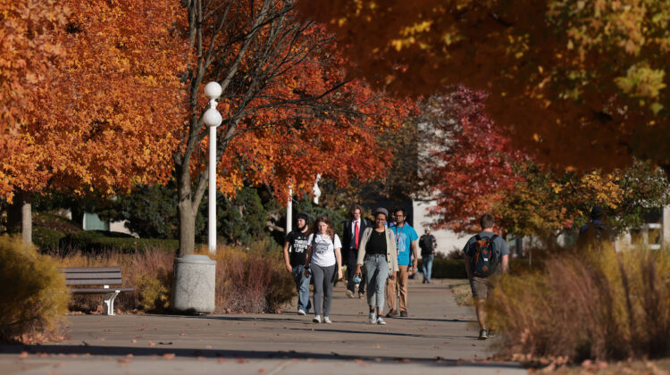 Students walk down campus path amid many brightly colored fall trees.
