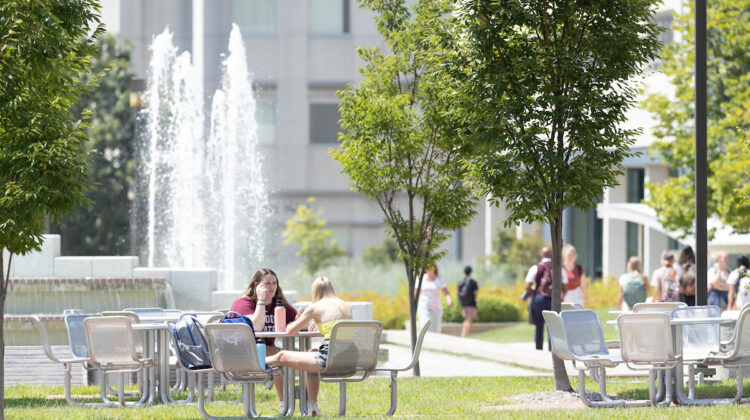 Students sit at picnic table with fountain in background.