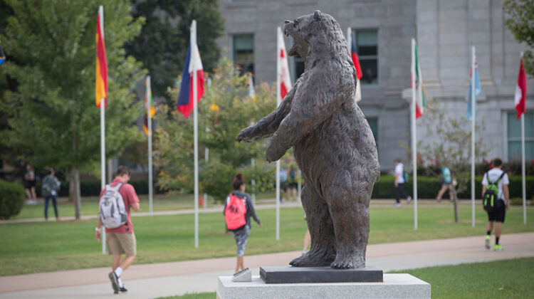 Students walk down the Avenue of Flags and pass the Bear statue on campus.