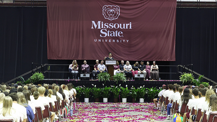 A graduation ceremony at Missouri State University features confetti on the floor, seated attendees, and a stage with a large banner and a screen displaying speakers.