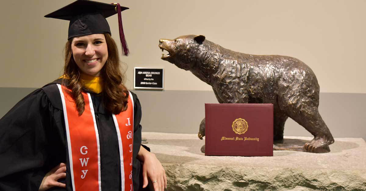 Kirby Williams wearing cap and gown and posing next to bear statue in JQH Arena on commencement day.